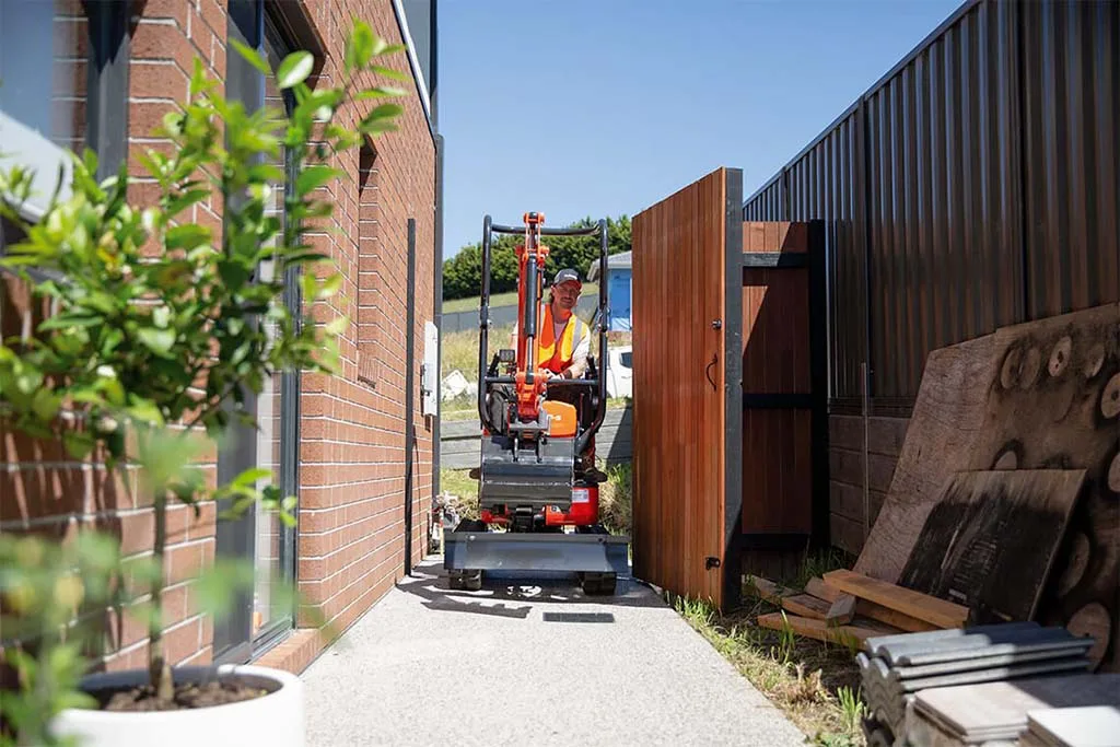 Kubota 1.2 tonne excavator driving through a side gate between a brick house and fence
