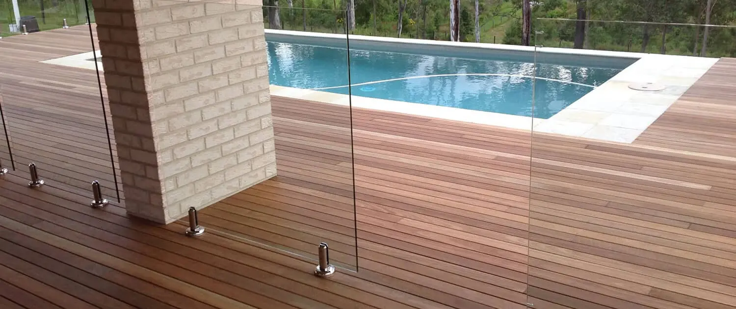 A modern outdoor space crafted by Builders Bayside Brisbane features a wooden deck with a glass railing overlooking a rectangular swimming pool. The pool area is surrounded by lush greenery and tall trees, creating a serene and inviting atmosphere. Builders Bayside Brisbane, Home Renovations Redlands