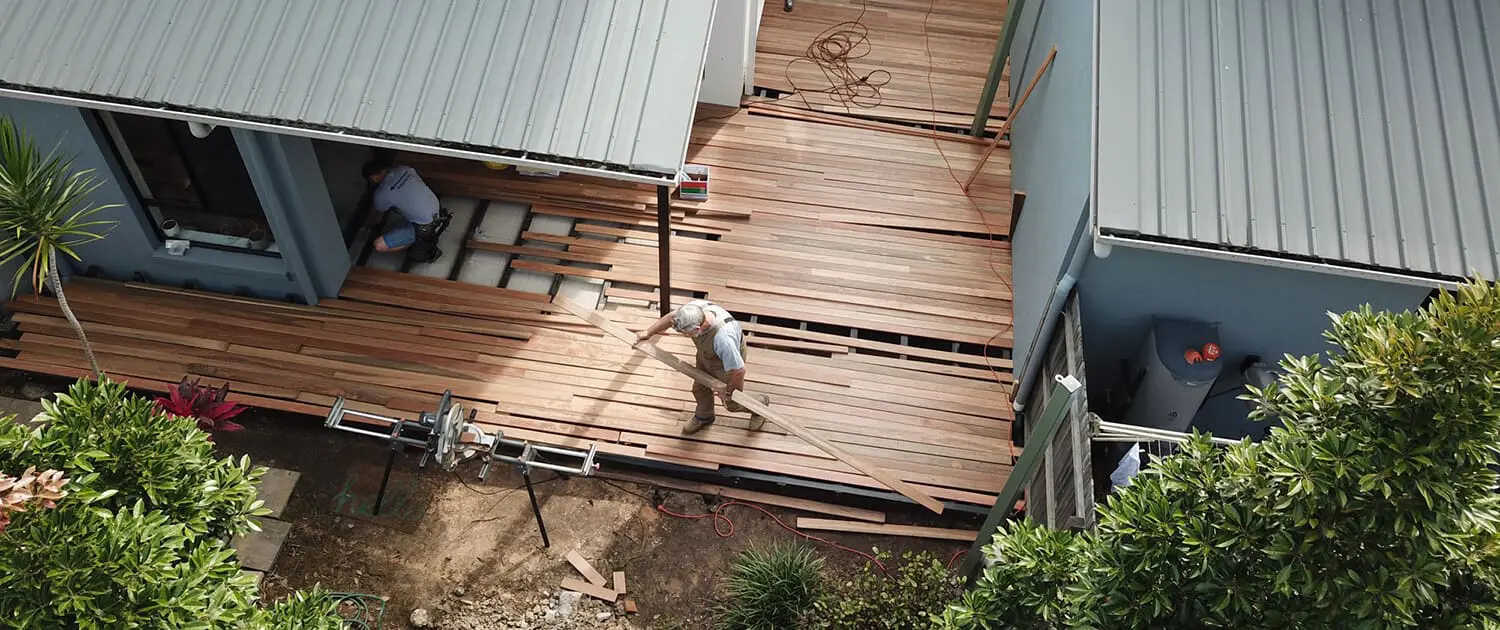 Aerial view of a construction site where wooden flooring is being installed outdoors. One person is working on the decking while another is partly visible under a structure. Various tools and materials are scattered around. The area is surrounded by greenery and buildings. Builders Bayside Brisbane, Home Renovations Redlands