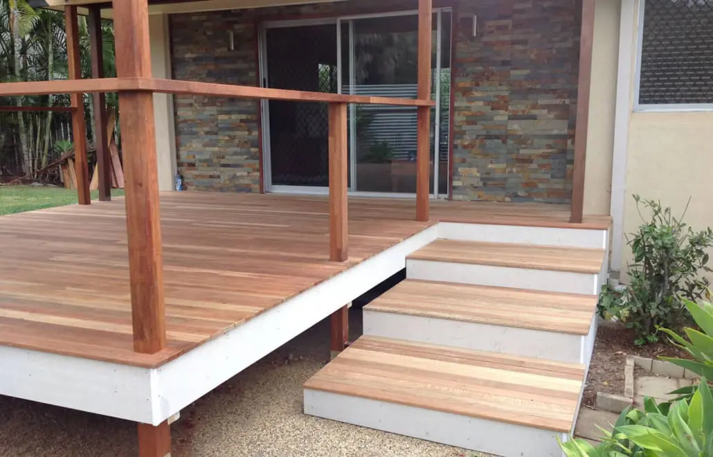 A backyard deck with wooden flooring and railings by Builders Bayside Brisbane leads up to a sliding glass door. There are three steps with a white base and wooden treads. The deck overlooks a garden area with lush green plants, and a stone accent wall is visible in the background. Builders Bayside Brisbane, Home Renovations Redlands