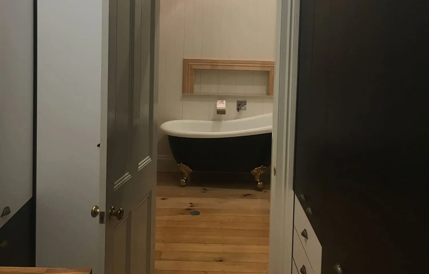 A narrow doorway opens into a bathroom featuring a vintage-style black clawfoot bathtub with gold feet. The tub is set against a white-paneled wall with a wooden shelf above it. The bathroom has natural wooden flooring and dark cabinetry on the right side. Builders Bayside Brisbane, Home Renovations Redlands