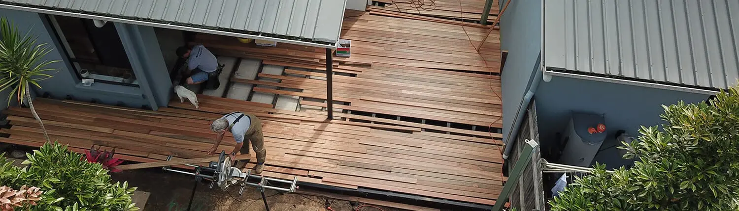 Aerial view of workers building a wooden deck between two blue buildings. One worker is using a saw, and another is laying planks, collaborating efficiently as part of Manttan Construction's skilled team. Tools and wooden planks are scattered around, with green leafy bushes and trees providing lush surroundings. Builders Bayside Brisbane, Home Renovations Redlands