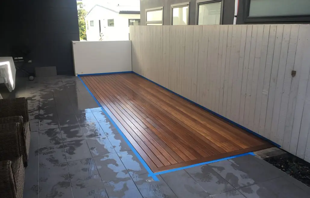 A narrow, rectangular wooden deck area is surrounded by gray tiles. Wet floor tiles can be seen on the left with wicker furniture and a grill. A white fence encloses the space, designed by Builders Bayside Brisbane, and modern buildings are visible in the background. Builders Bayside Brisbane, Home Renovations Redlands