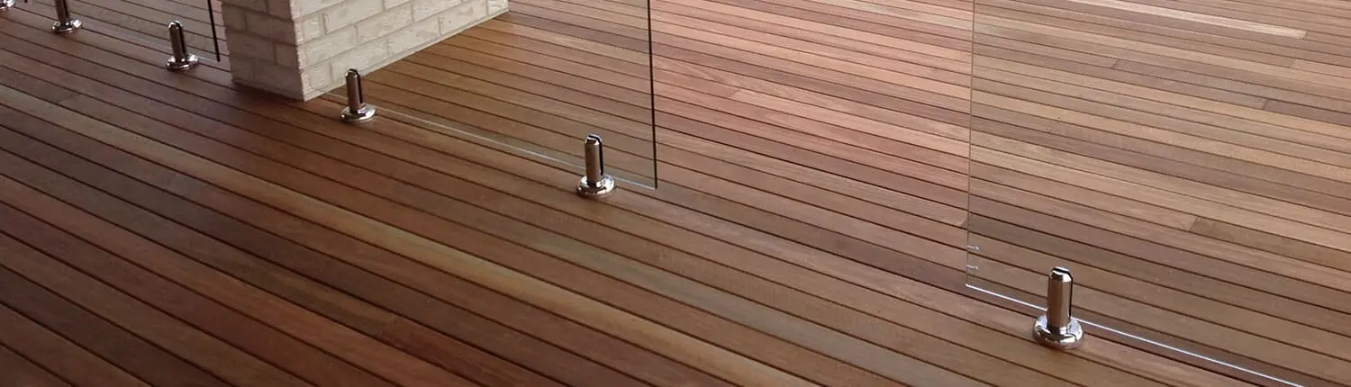 A wooden deck area with sleek planks in various shades of brown, crafted by Manttan Construction. The deck is divided by a clear glass railing, supported by modern stainless-steel brackets. A brick pillar is visible near the glass railing, adding a contrasting texture to the scene, perfect for home renovations in Redlands. Builders Bayside Brisbane, Home Renovations Redlands