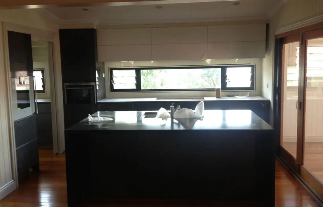 A modern kitchen with an island counter featuring a dark, polished surface. Behind the island is a row of cabinets and a large horizontal window, allowing natural light to filter in. The floor has a warm wooden finish, and sliding glass doors are visible on the right, crafted by Builders Bayside Brisbane. Builders Bayside Brisbane, Home Renovations Redlands