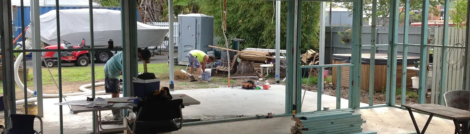 Construction site with two workers in a partially built structure. The frame of the building is visible, along with scattered tools and materials. One worker is kneeling, and the other is standing. Outside, there are vehicles and additional construction materials from Builders Bayside Brisbane. Builders Bayside Brisbane, Home Renovations Redlands