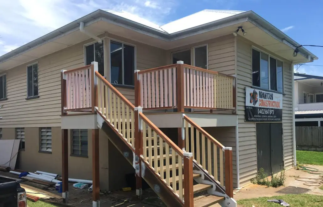 A two-story house with beige siding and a white roof is shown. The house, crafted by Manttan Construction, features a wooden exterior staircase leading to a small porch with railings in matching beige and brown colors. There is a construction sign on the side of the house, and some materials are nearby. Builders Bayside Brisbane, Home Renovations Redlands