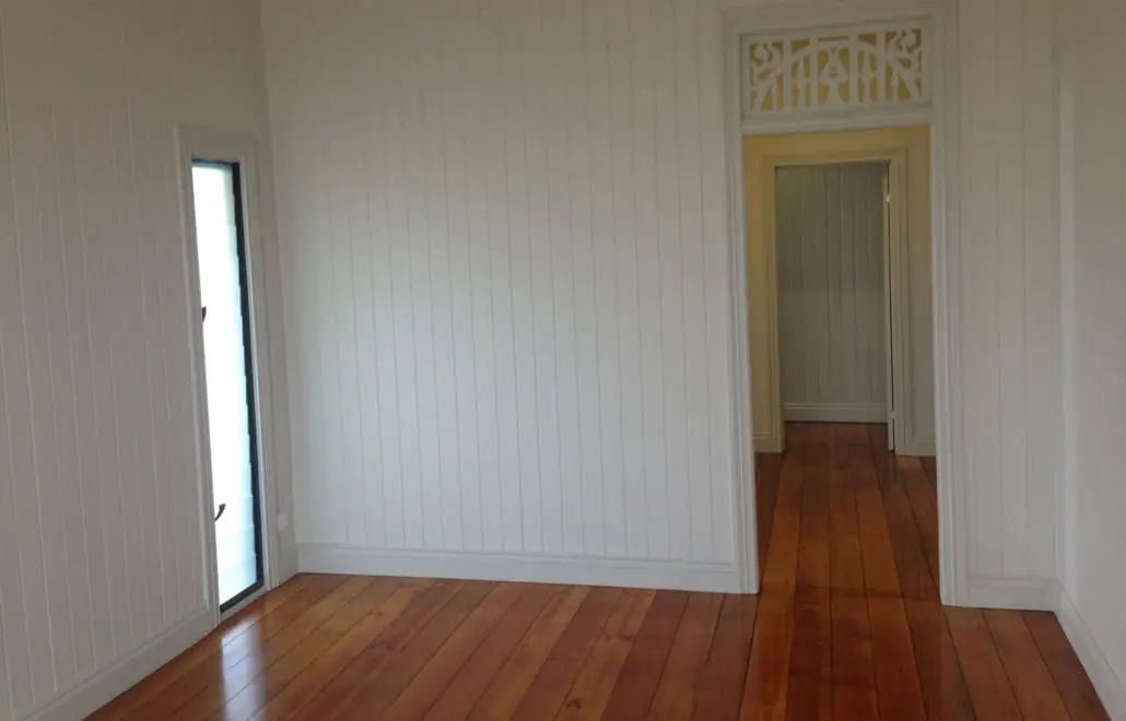 An empty room with wooden floors, white vertical wood-paneled walls, and a small vertical window on the left. A doorway leads to another room with similar flooring and a decorative woodwork detail above the door, showcasing the craftsmanship of Manttan Construction. Builders Bayside Brisbane, Home Renovations Redlands